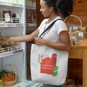 Woman carrying a shopping bag with a graphic representation of a red barn and leaves. Includes the text: Farmer's Market.