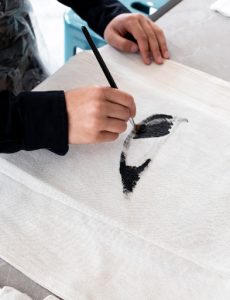 Artist's hands shown painting a design on a book bag.